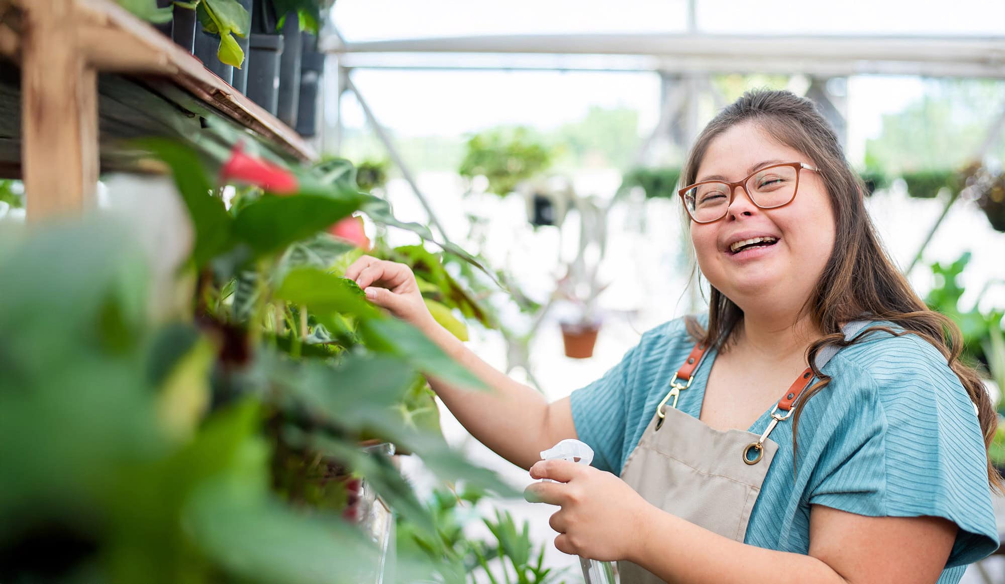 A woman wearing glasses and an apron smiles while watering plants in a greenhouse, reflecting support for Louisiana citizens with developmental disabilities.