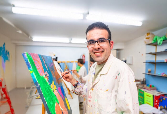 A man wearing glasses and a paint-splattered smock smiles while painting, supported by the Louisiana Developmental Disabilities Council.