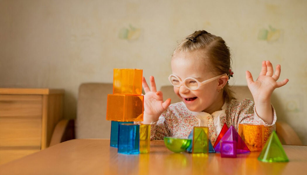 A young girl with glasses smiles and raises her hands, playing with blocks—a joyful moment supported by the Louisiana Developmental Disabilities Council.