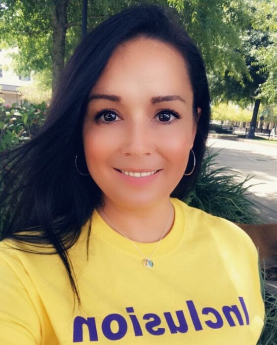 A woman with long dark hair smiles outside in a yellow “Inclusion” shirt, showing support for Louisiana citizens with developmental disabilities.