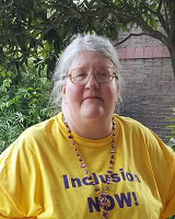 An older woman in glasses wears a yellow “Inclusion NOW!” t-shirt, showing support for Louisiana citizens with developmental disabilities.