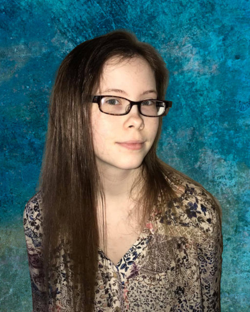 A young person with long brown hair and glasses poses in front of a blue background, showing support for Louisiana citizens with developmental disabilities.