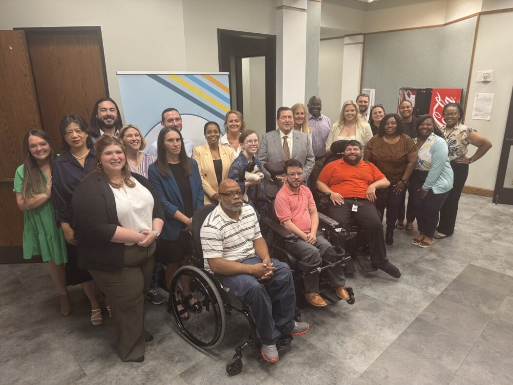 A group of people, including individuals in wheelchairs, pose together and smile for a photo in an office hallway.
