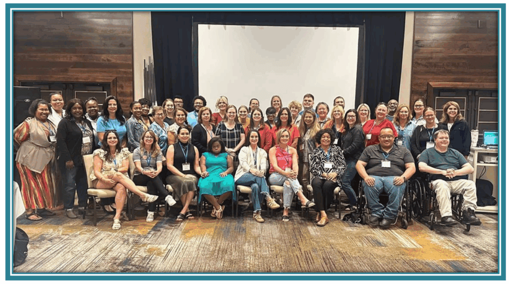 A large group of people pose together indoors in front of a projection screen, with some seated and others standing.