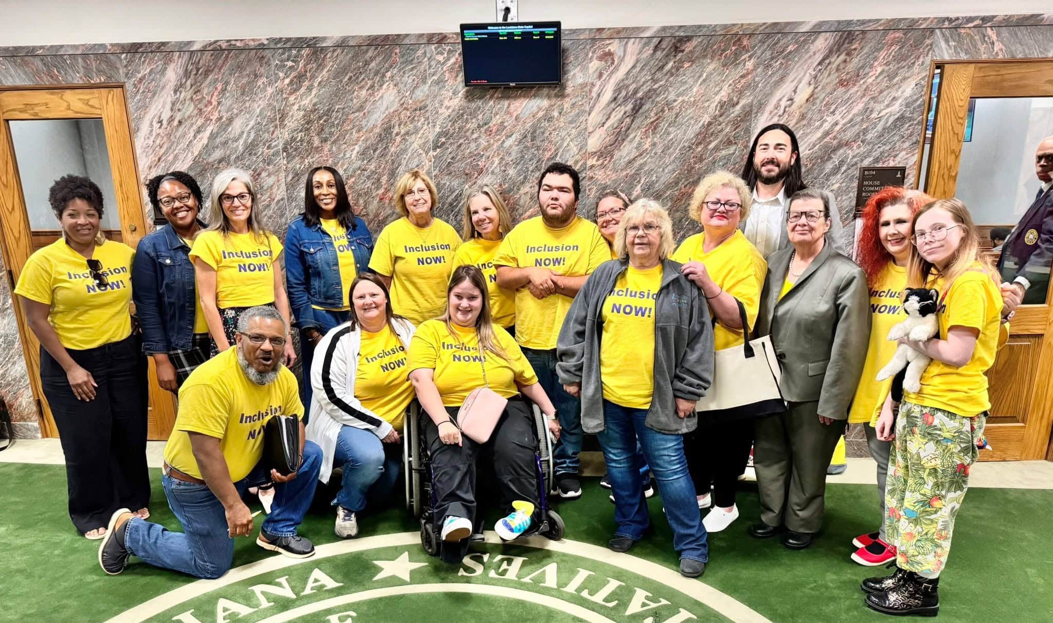 A group of people, many in yellow "Inclusion NOW!" shirts, pose for a photo together inside a government building.